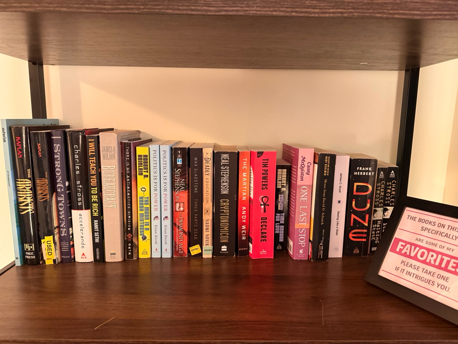 A bookshelf of Dave Kasten’s favorite books next to a sign reading “The books on this shelf specifically are some of my favorites. Please take one if it intrigues you.”